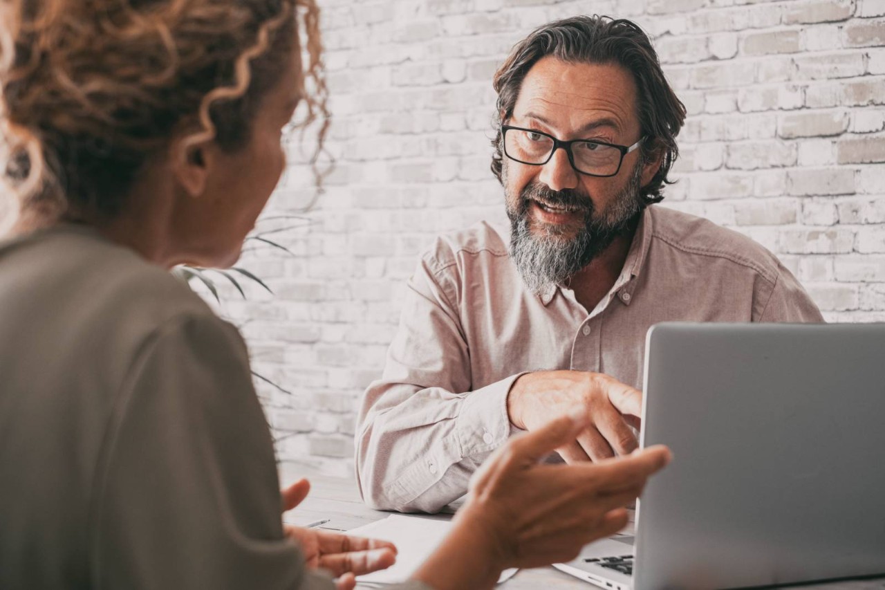 Person pointing at a laptop whilst speaking to another person, both sitting opposite sides of a desk